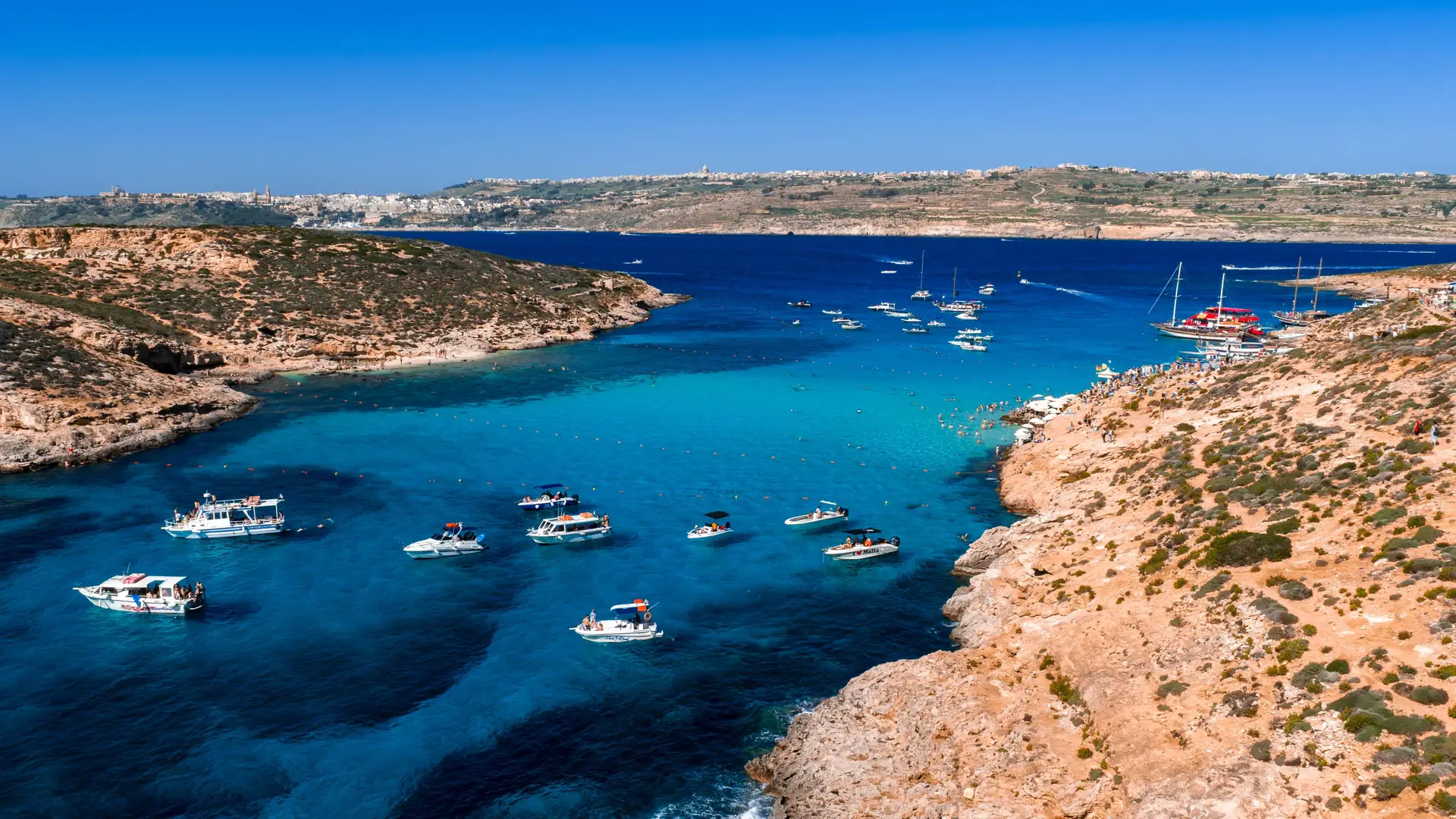 Boat tours Malta: aerial view of motorboats and yachts anchored in a stunning turquoise cove with golden cliffs and sandy beach